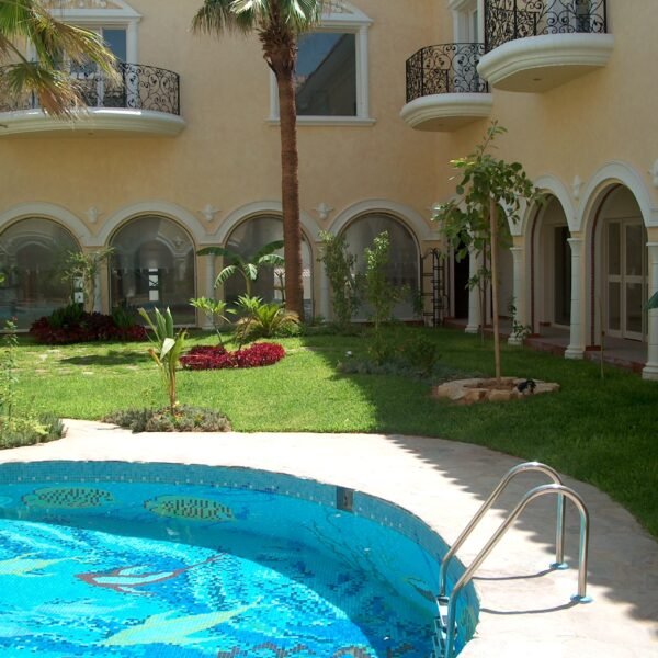 مسبح دائري مع درجات دخول وحديقة مزروعة بأشجار ونباتات متنوعة.