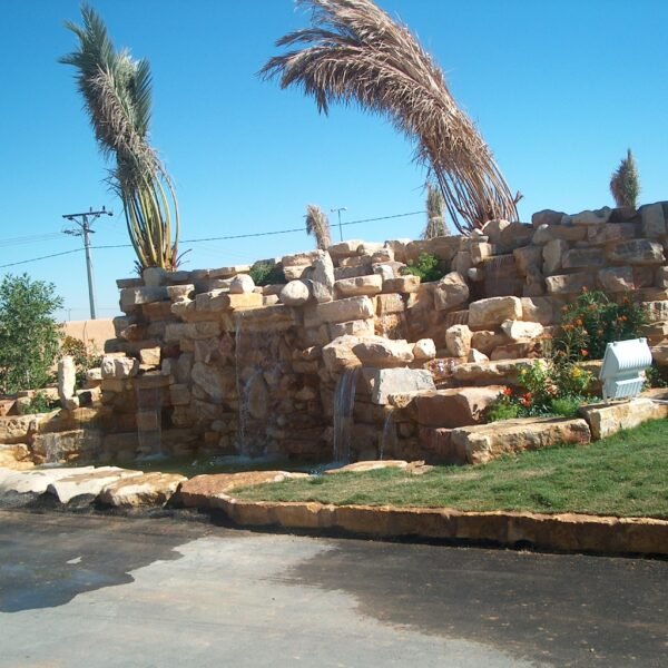 شلال صناعي يتكون من صخور كبيرة مع نباتات خضراء وأشجار نخيل في الخلفية.