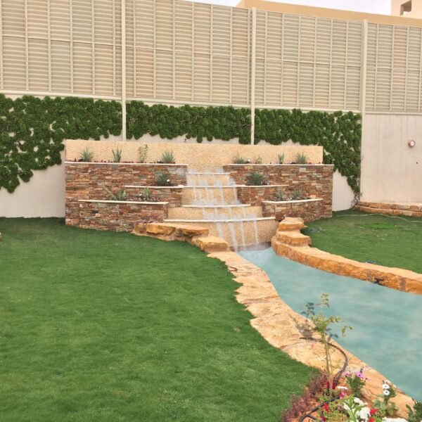 شلال مائي يتدفق من جدار حجري في حديقة مع نباتات خضراء وأرضية عشبية.