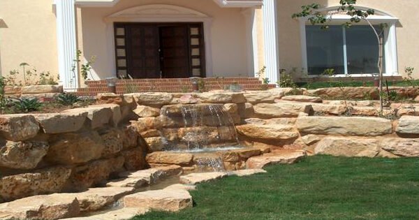شلال مياه يتدفق من الصخور الطبيعية في حديقة منزلية مع واجهة منزلية خلفية.